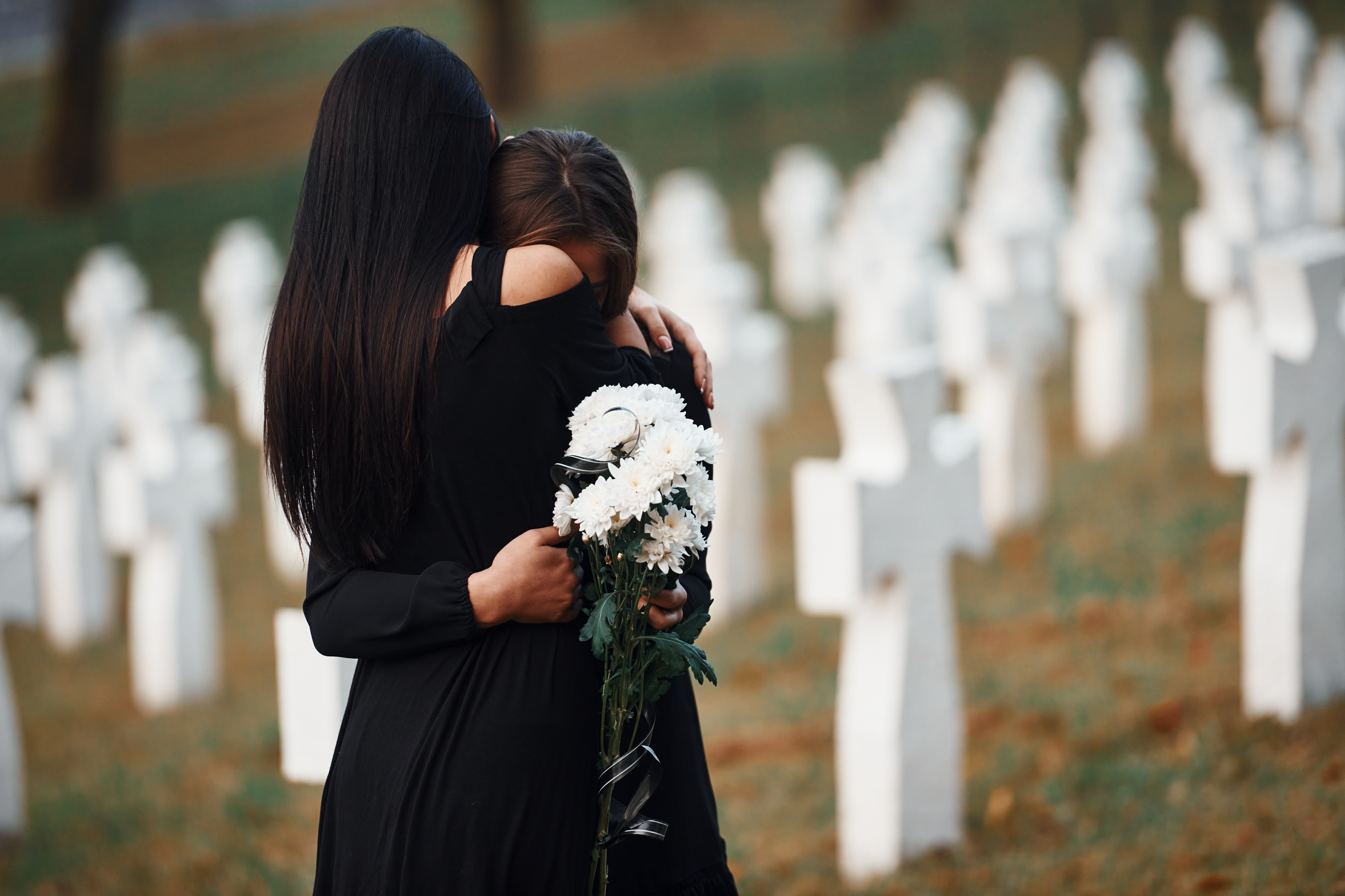 Embracing each other and crying. Two young women in black clothes visiting cemetery with many white crosses
