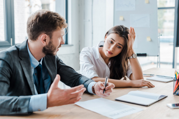 Two people at a desk discussing paperwork. One looks frustrated.